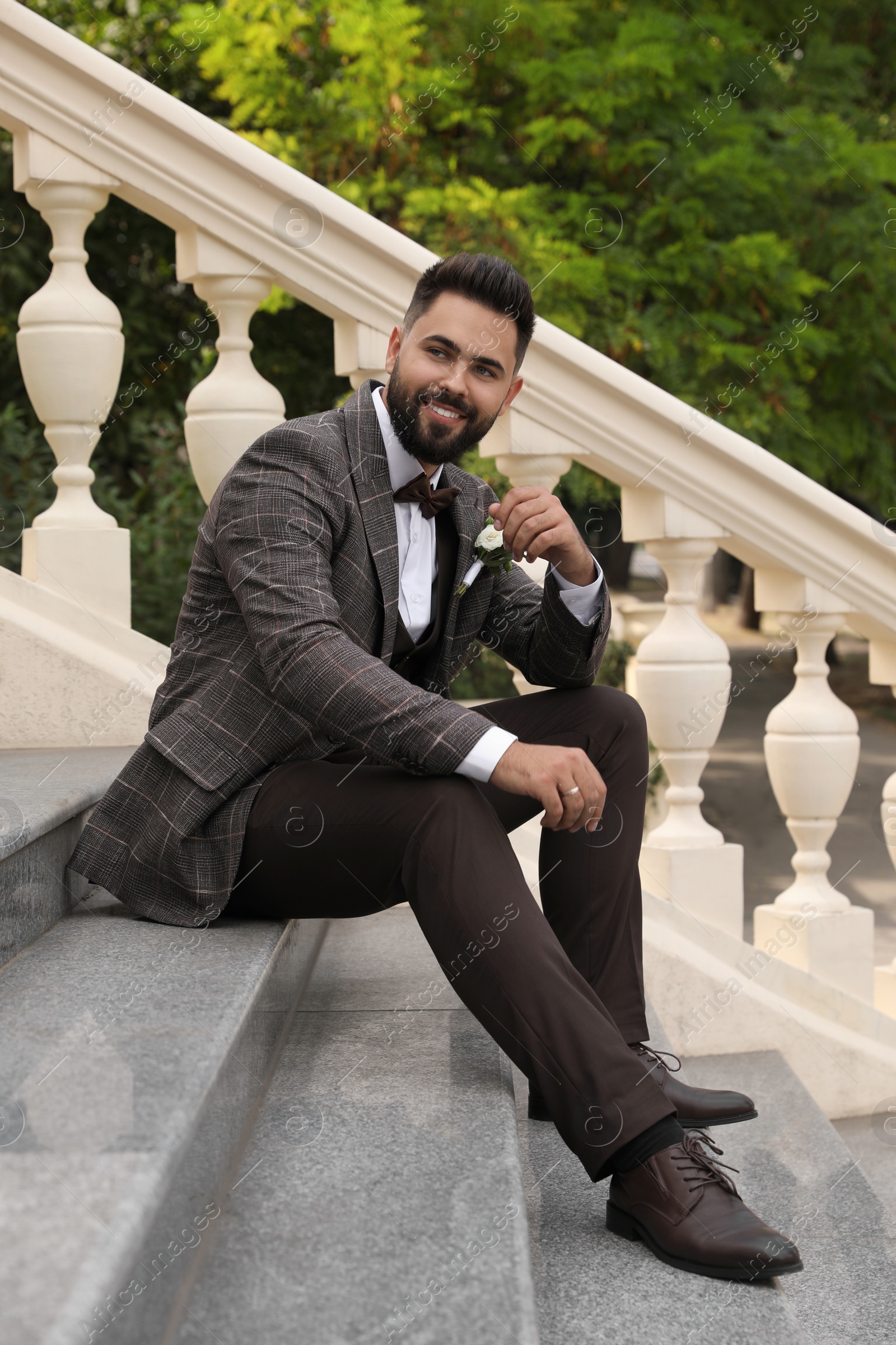 Photo of Young groom in elegant suit with beautiful boutonniere sitting on stairs outdoors