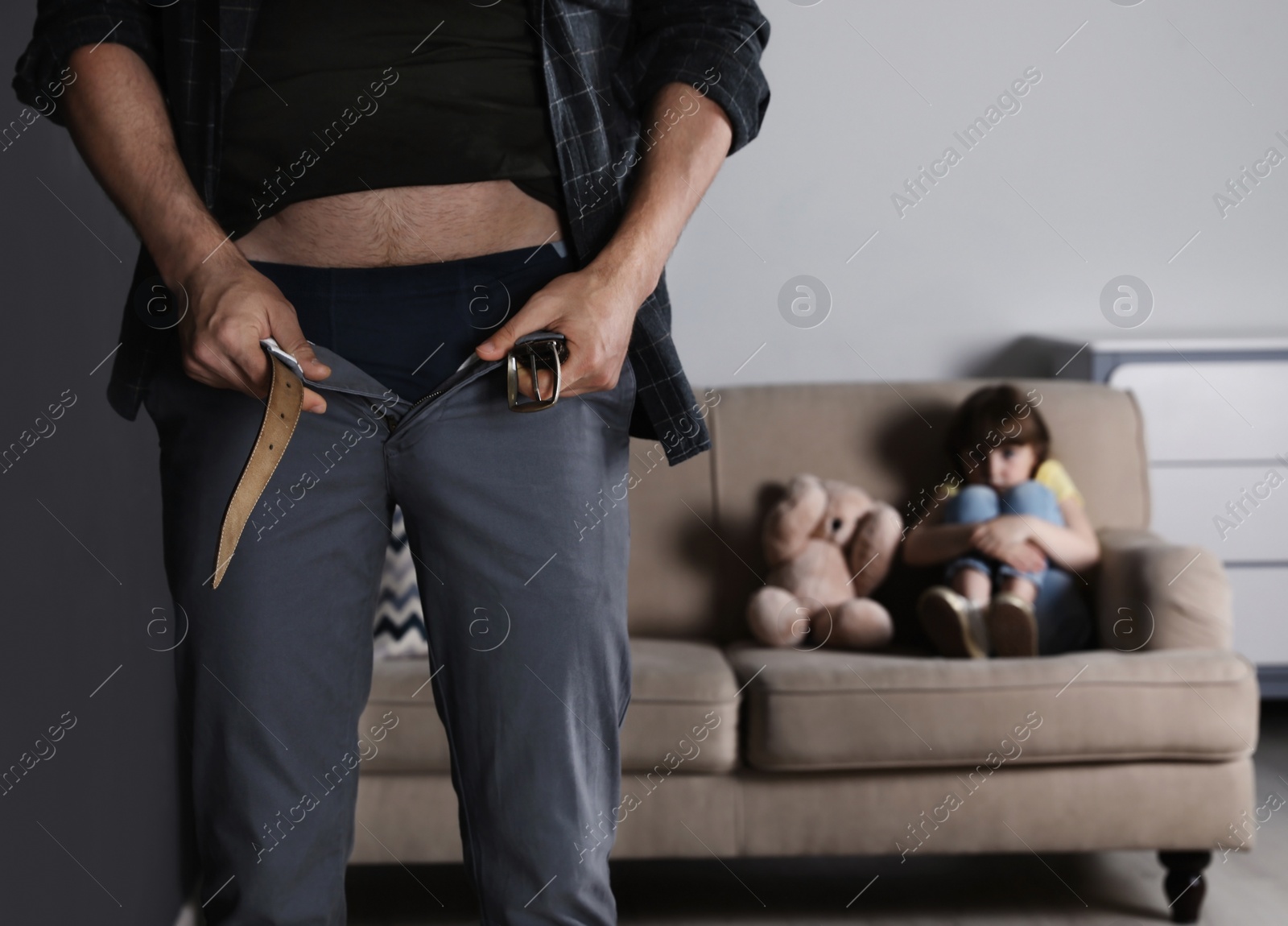 Photo of Man with unzipped pants standing near scared little girl on sofa indoors. Child in danger