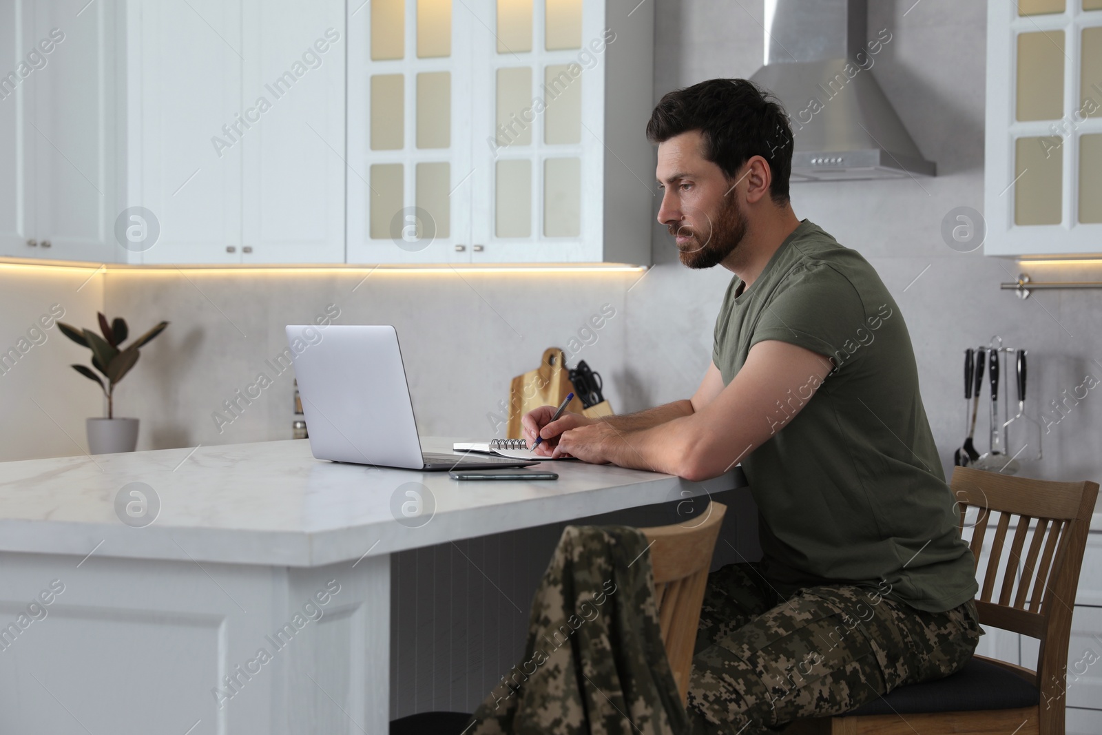 Photo of Soldier taking notes while working with laptop at white marble table in kitchen. Military service