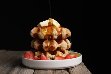 Photo of Pouring honey onto delicious Belgian waffles with strawberries and butter at wooden table against black background, space for text
