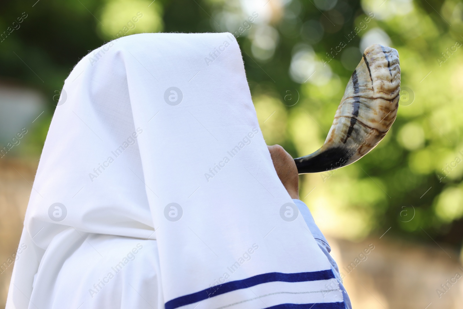Photo of Jewish man in tallit blowing shofar outdoors. Rosh Hashanah celebration