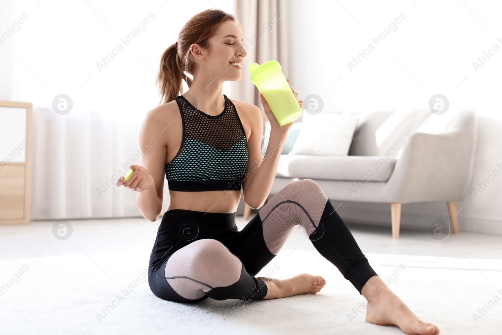 Photo of Young sporty woman with bottle of protein shake sitting on floor at home