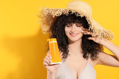 Photo of Beautiful young woman in straw hat holding tube of sunscreen against orange background, space for text