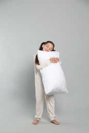 Cute girl in white pajamas hugging pillow on light grey background