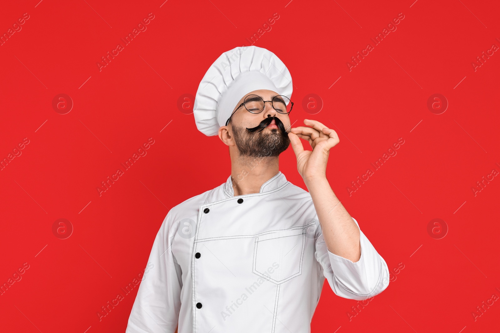 Photo of Professional chef with funny artificial moustache showing perfect sign on red background