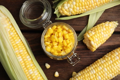 Photo of Tasty sweet corn kernels in jar and fresh cobs on wooden table, flat lay