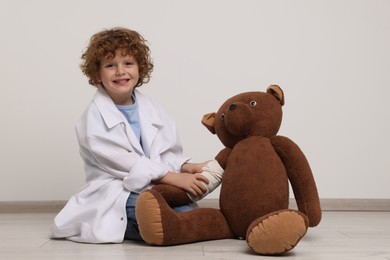 Photo of Little boy in medical uniform and toy bear with bandage indoors