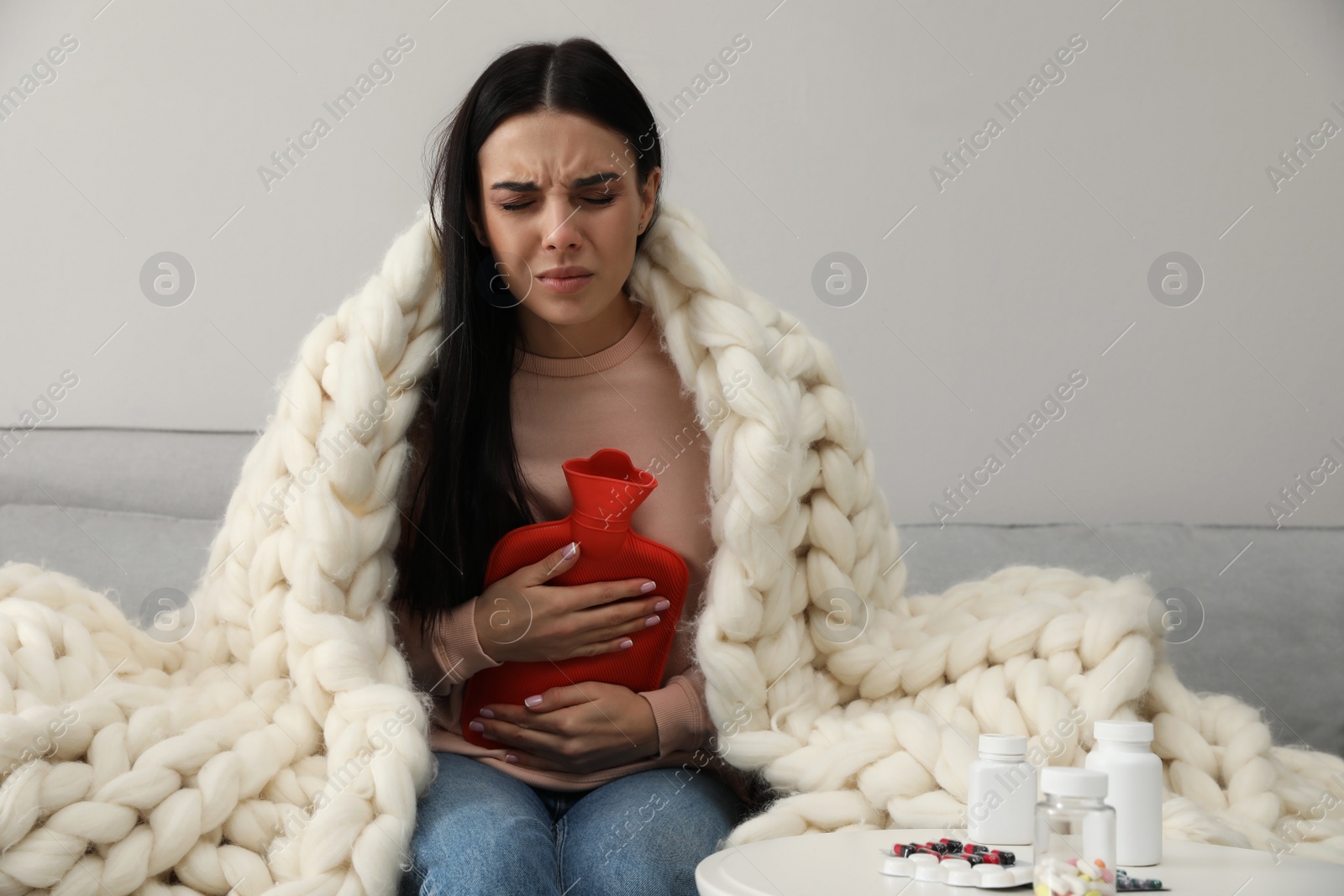 Photo of Woman using hot water bottle to relieve abdominal pain on sofa at home
