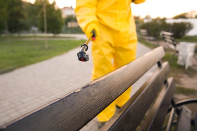 Photo of Person in hazmat suit disinfecting bench in park with sprayer, closeup. Surface treatment during coronavirus pandemic
