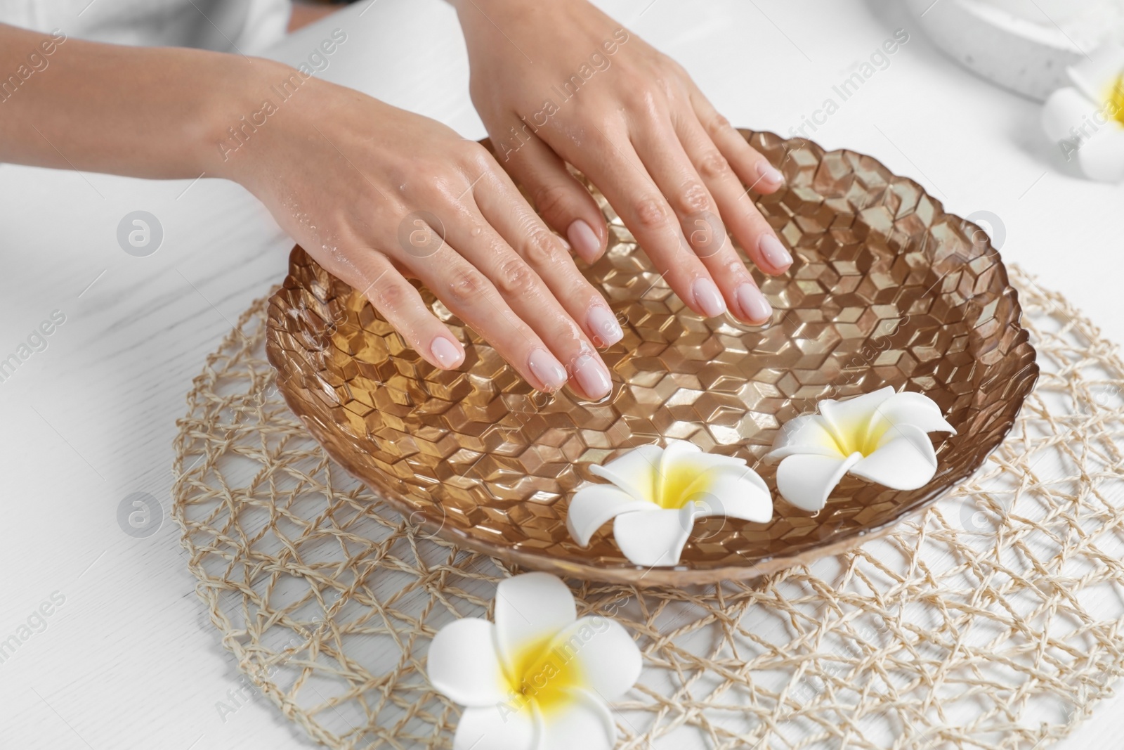 Photo of Woman soaking her hands in bowl with water and flowers on table, closeup. Spa treatment