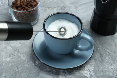 Photo of Whisking milk in cup with mini mixer (milk frother) on grey textured table, closeup