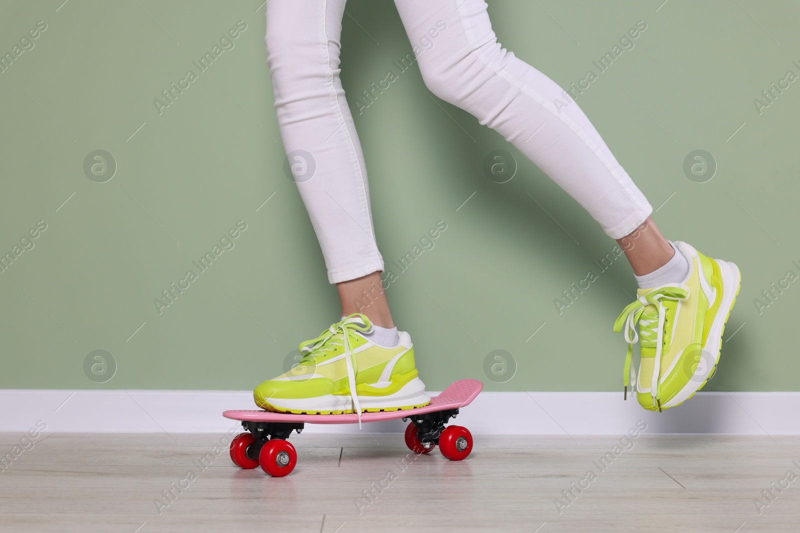 Photo of Woman in new stylish sneakers standing on skateboard near light green wall, closeup