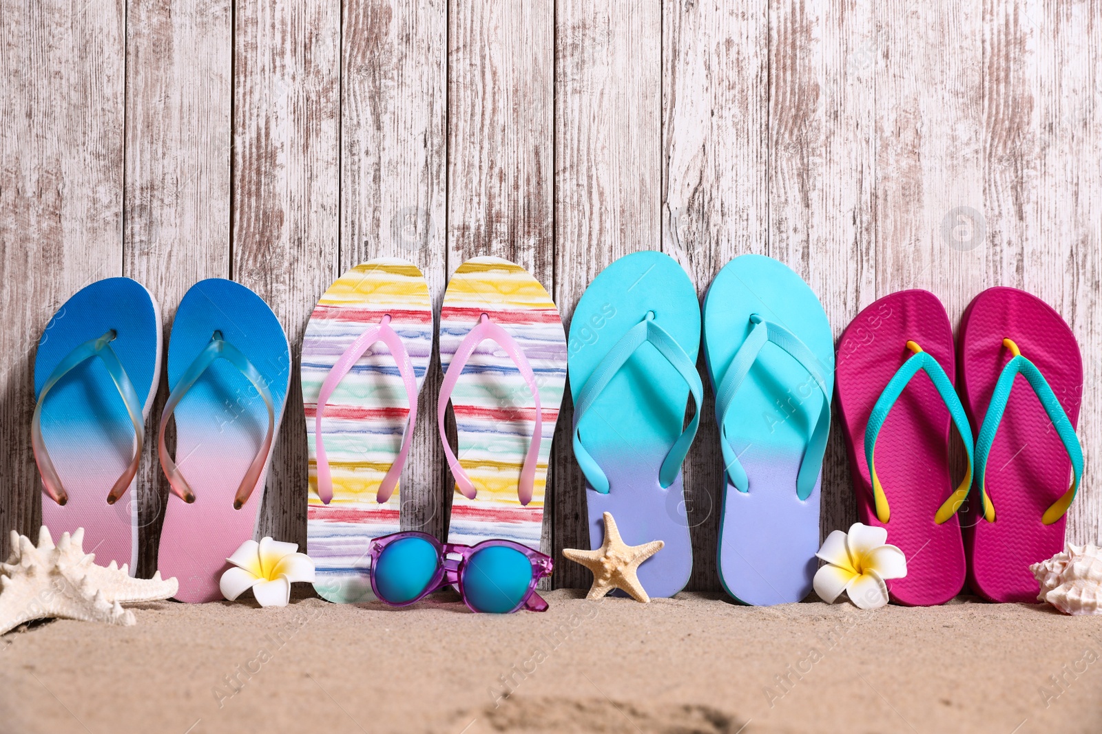 Photo of Different bright flip flops and sunglasses on sand near wooden wall, space for text. Summer beach accessories