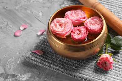 Photo of Tibetan singing bowl with water, beautiful roses and mallet on grey textured table, space for text