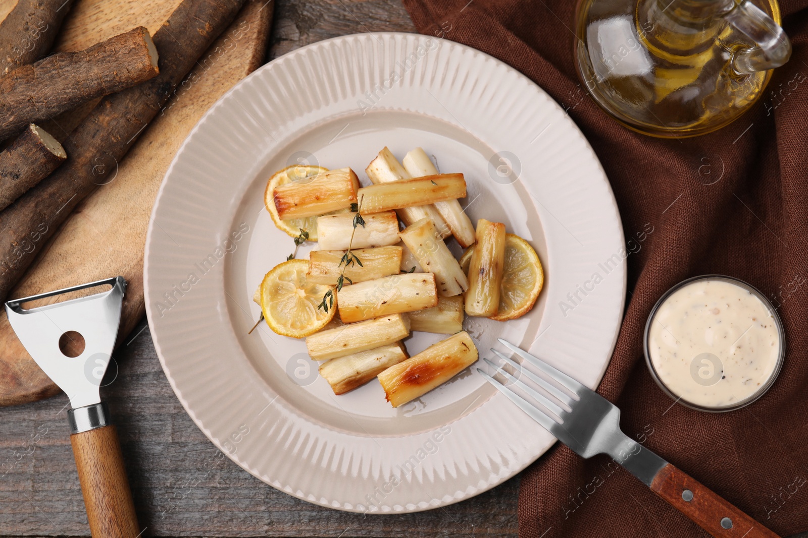 Photo of Plate with baked salsify roots, lemon, fork and sauce on wooden table, flat lay