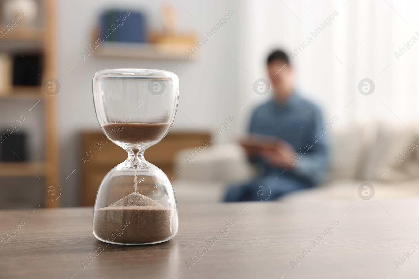 Photo of Hourglass with flowing sand on desk. Man reading book in room, selective focus