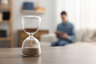 Hourglass with flowing sand on desk. Man reading book in room, selective focus