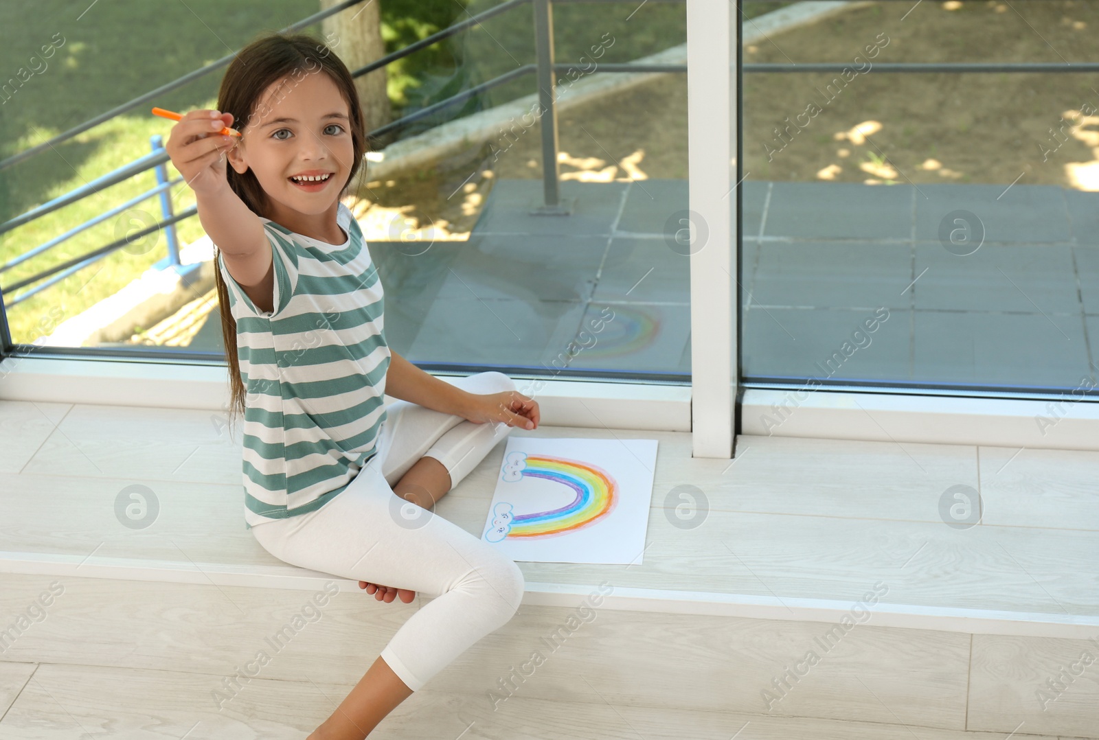 Photo of Little girl drawing rainbow near window indoors. Stay at home concept