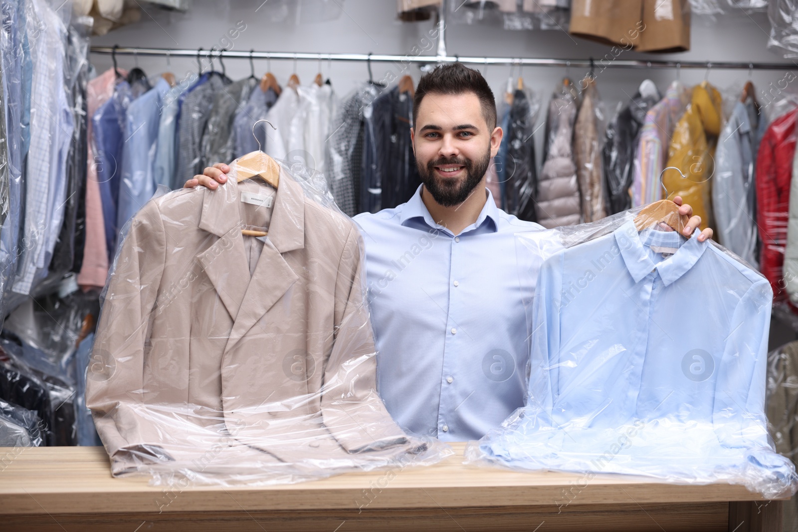 Photo of Dry-cleaning service. Happy worker holding hangers with clothes in plastic bags at counter indoors