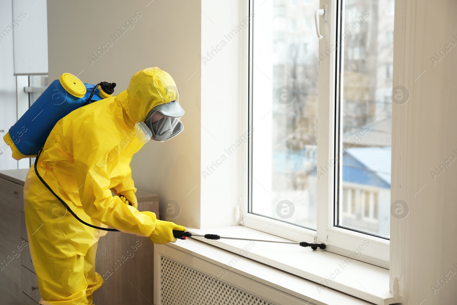 Photo of Pest control worker in protective suit spraying pesticide near window indoors