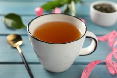 Photo of Cup of herbal diet tea and measuring tape on light blue wooden table, closeup. Weight loss concept