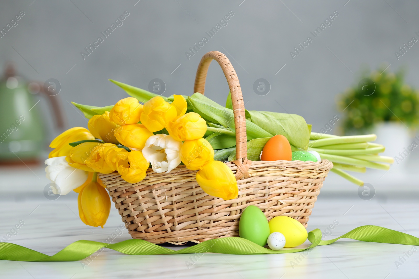 Photo of Easter basket with bouquet of tulips and eggs on white marble table indoors