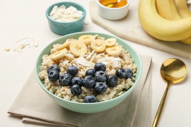 Photo of Tasty oatmeal with banana, blueberries, coconut flakes and honey served in bowl on beige table