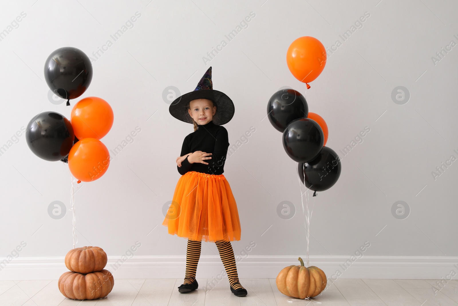 Photo of Cute little girl with balloons and pumpkins wearing Halloween costume near light wall