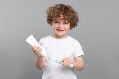 Photo of Cute little boy squeezing toothpaste from tube onto electric toothbrush on light grey background