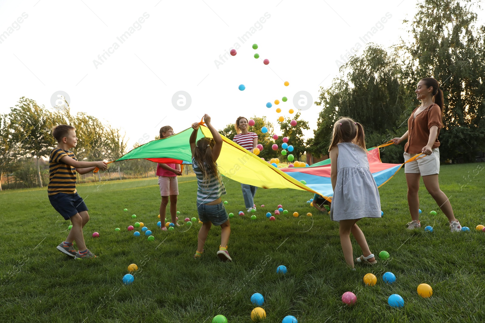 Photo of Group of children and teachers playing with rainbow playground parachute on green grass. Summer camp activity