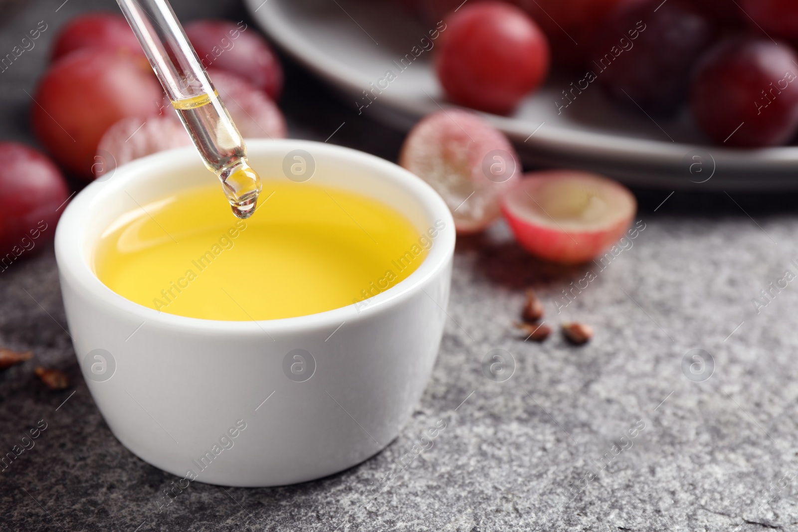 Photo of Dripping natural grape seed oil into bowl on grey table. Organic cosmetic