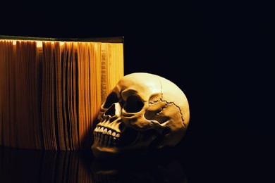 Photo of Human skull and old book on mirror table against black background. Space for text