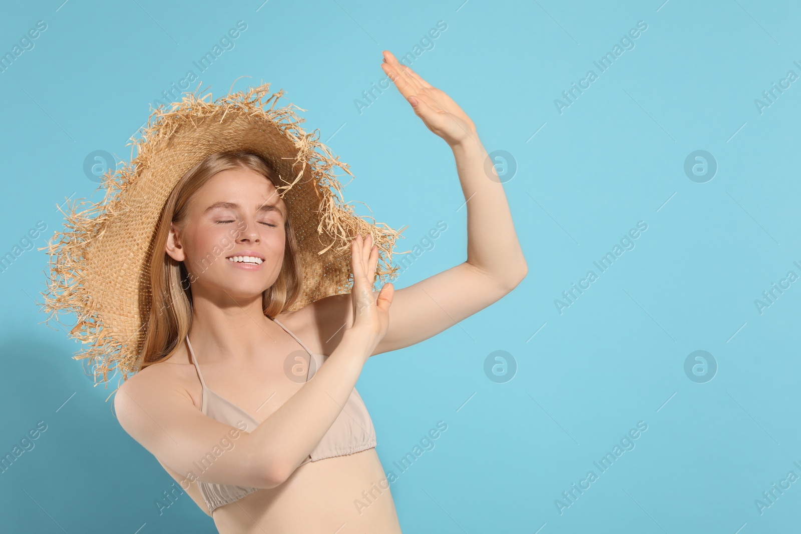 Photo of Beautiful young woman in straw hat shading herself with hands from sunlight on light blue background, space for text