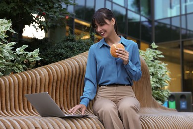 Photo of Lunch time. Happy businesswoman with hamburger using laptop on bench outdoors