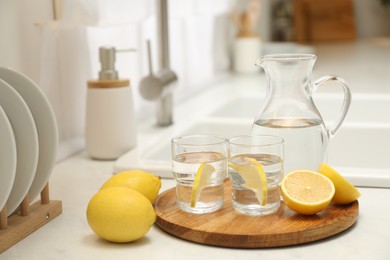 Jug, glasses with clear water and lemons on white table in kitchen