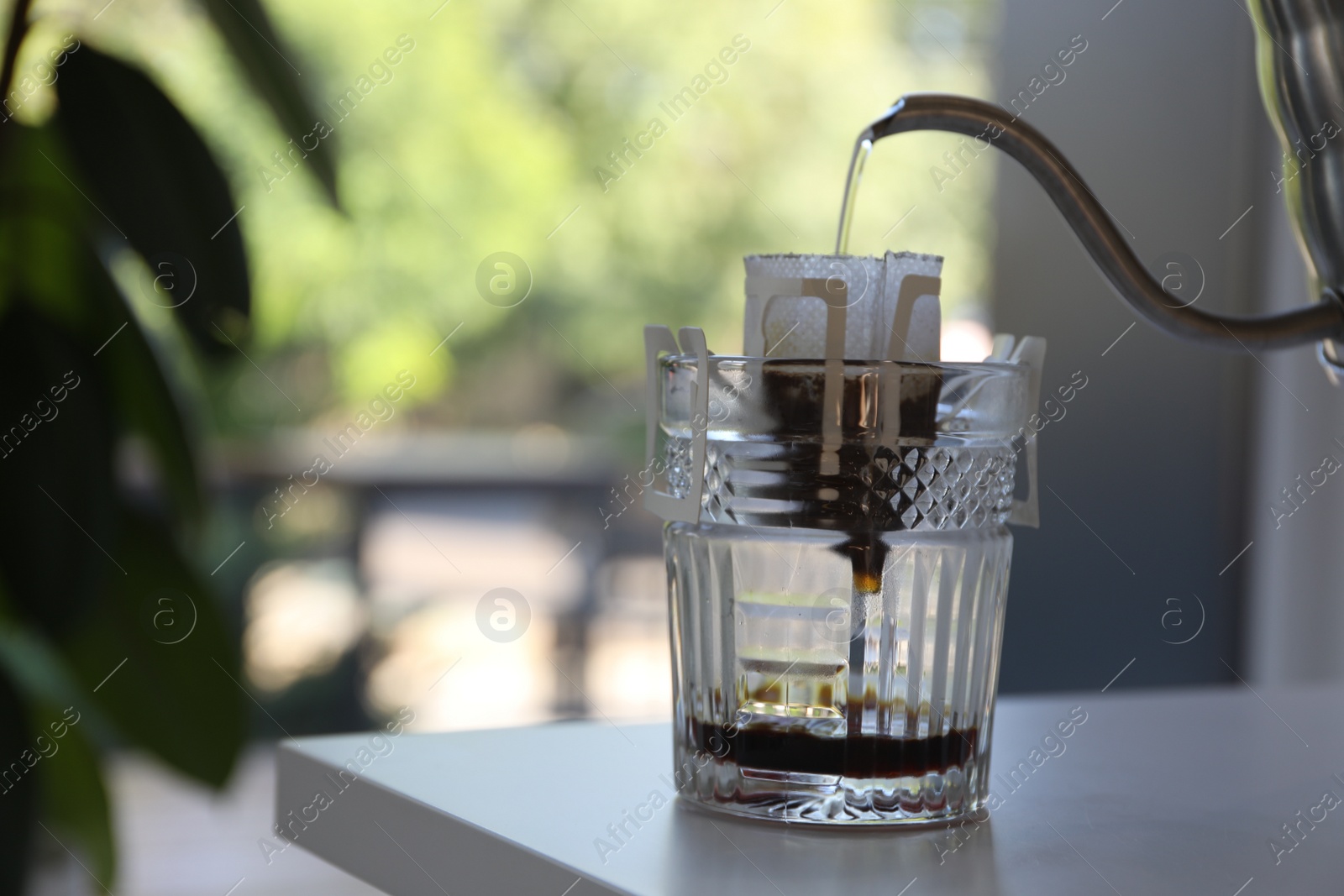 Photo of Pouring hot water into glass with drip coffee bag from kettle on white table, closeup. Space for text