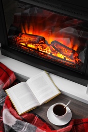 Cup of hot coffee, plaid and book on floor near fireplace, above view. Cozy atmosphere