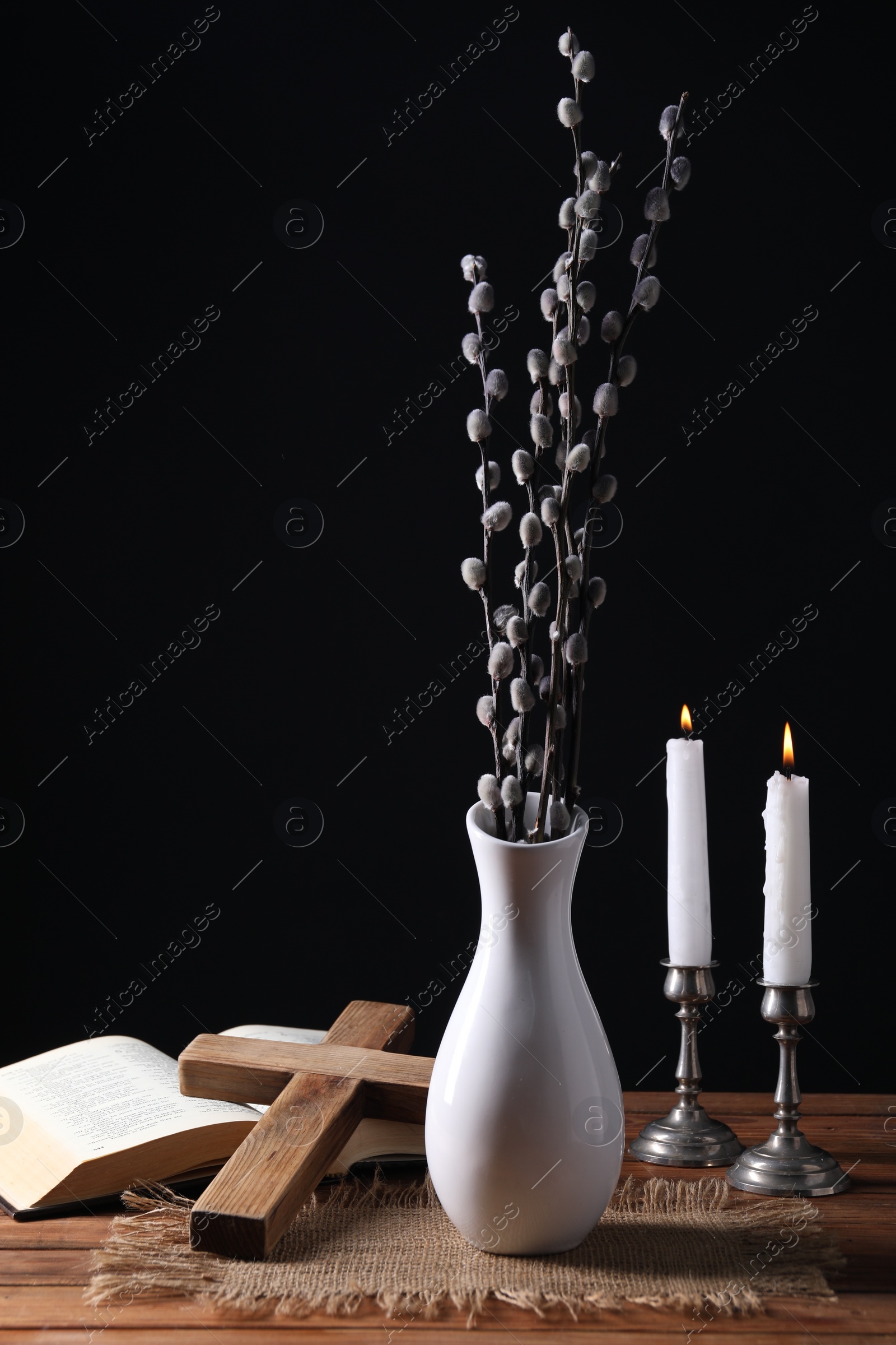 Photo of Burning church candles, cross, Bible and vase of willow branches on wooden table