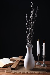 Burning church candles, cross, Bible and vase of willow branches on wooden table