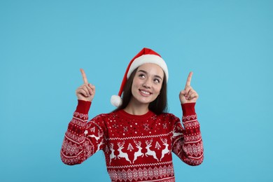 Photo of Happy young woman in Christmas sweater and Santa hat pointing at something on light blue background