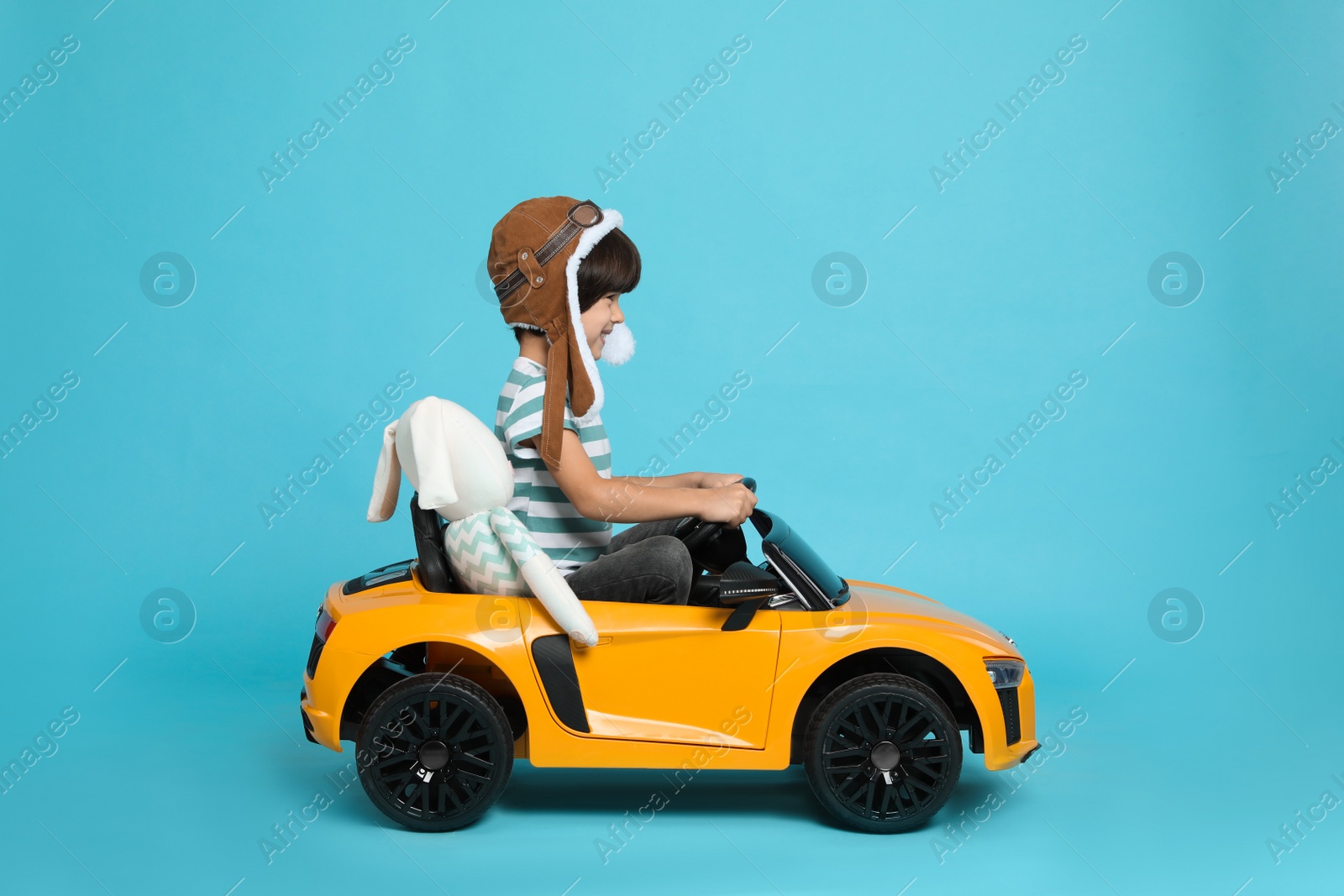 Photo of Cute little boy with toy bunny driving children's car on light blue background