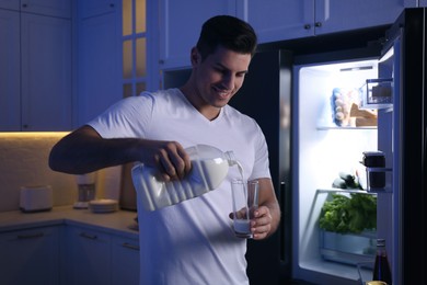 Man pouring milk from gallon bottle into glass near refrigerator in kitchen at night