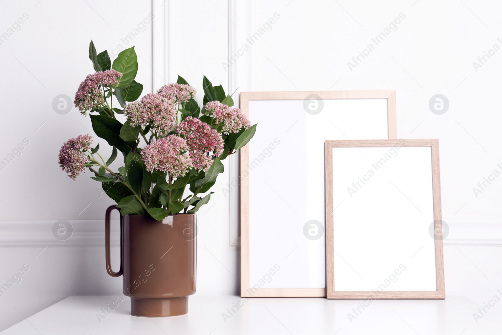 Photo of Stylish ceramic vase with beautiful flowers and blank frames on table near white wall
