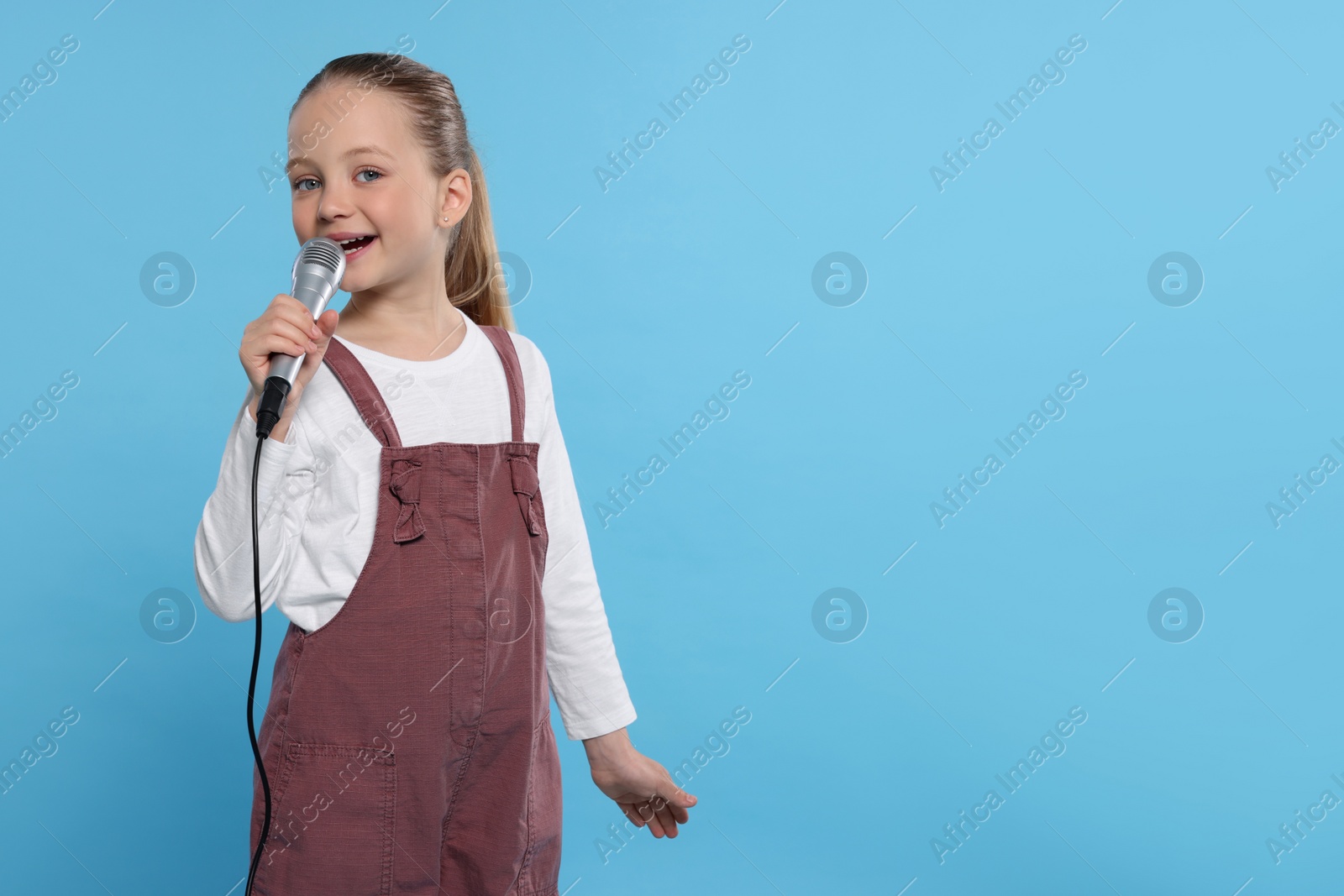 Photo of Cute little girl with microphone singing on light blue background, space for text