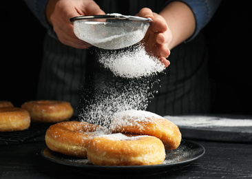 Photo of Woman decorating delicious donuts with powdered sugar at black table, closeup