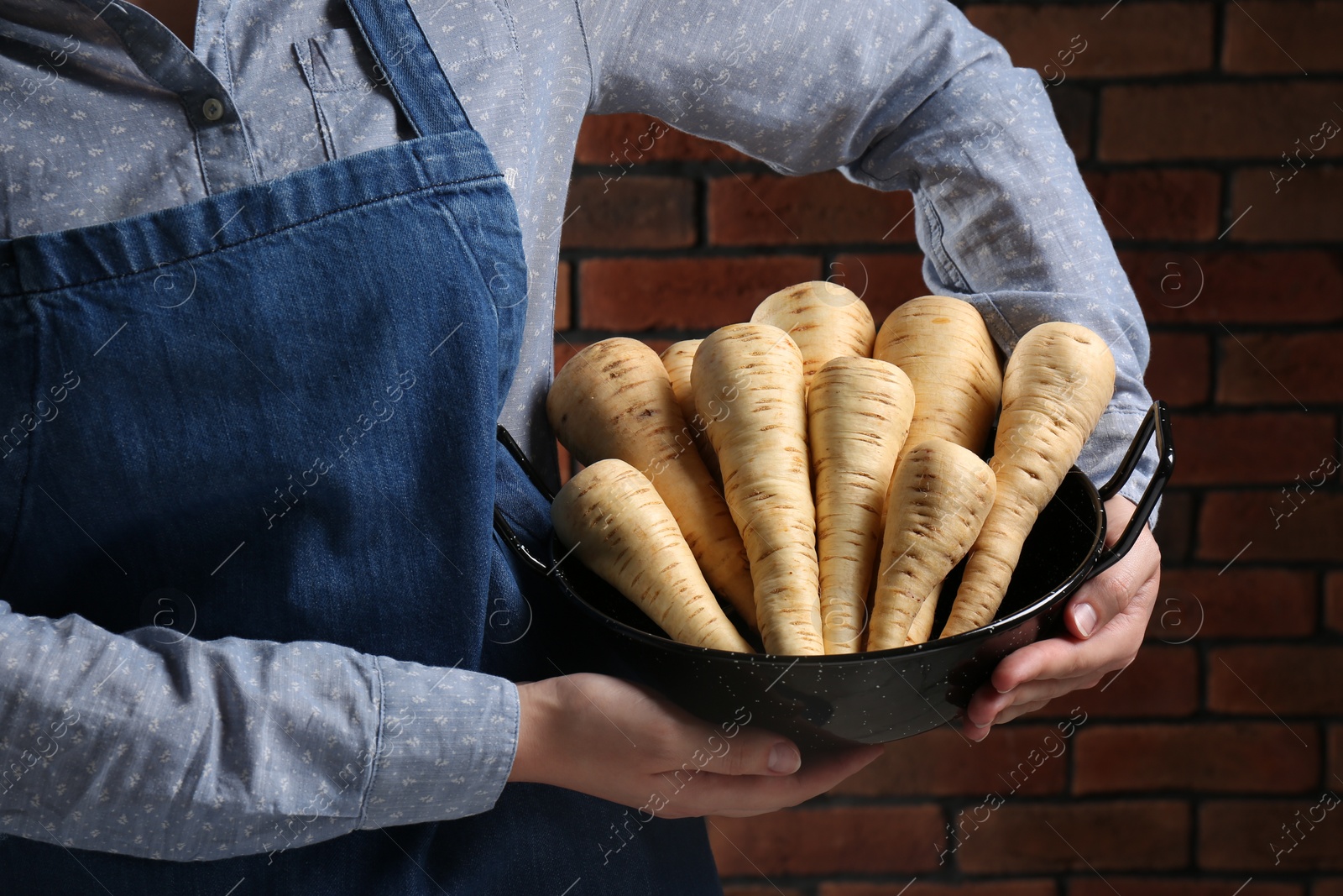 Photo of Woman holding bowl with fresh ripe parsnips near red brick wall, closeup