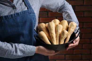 Woman holding bowl with fresh ripe parsnips near red brick wall, closeup