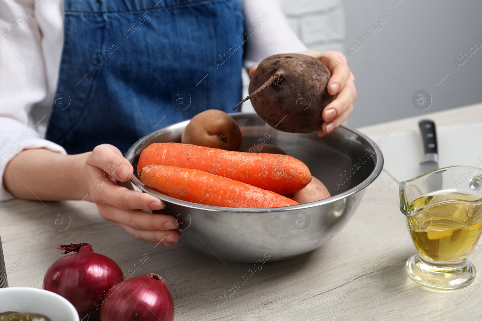 Photo of Woman putting beet into bowl with fresh vegetables at white wooden table, closeup. Cooking vinaigrette salad