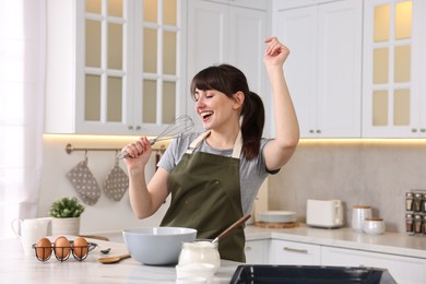 Happy young housewife with whisk having fun while cooking at white marble table in kitchen