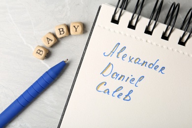 Photo of Notebook with different baby names, pen and wooden cubes on grey marble table, flat lay
